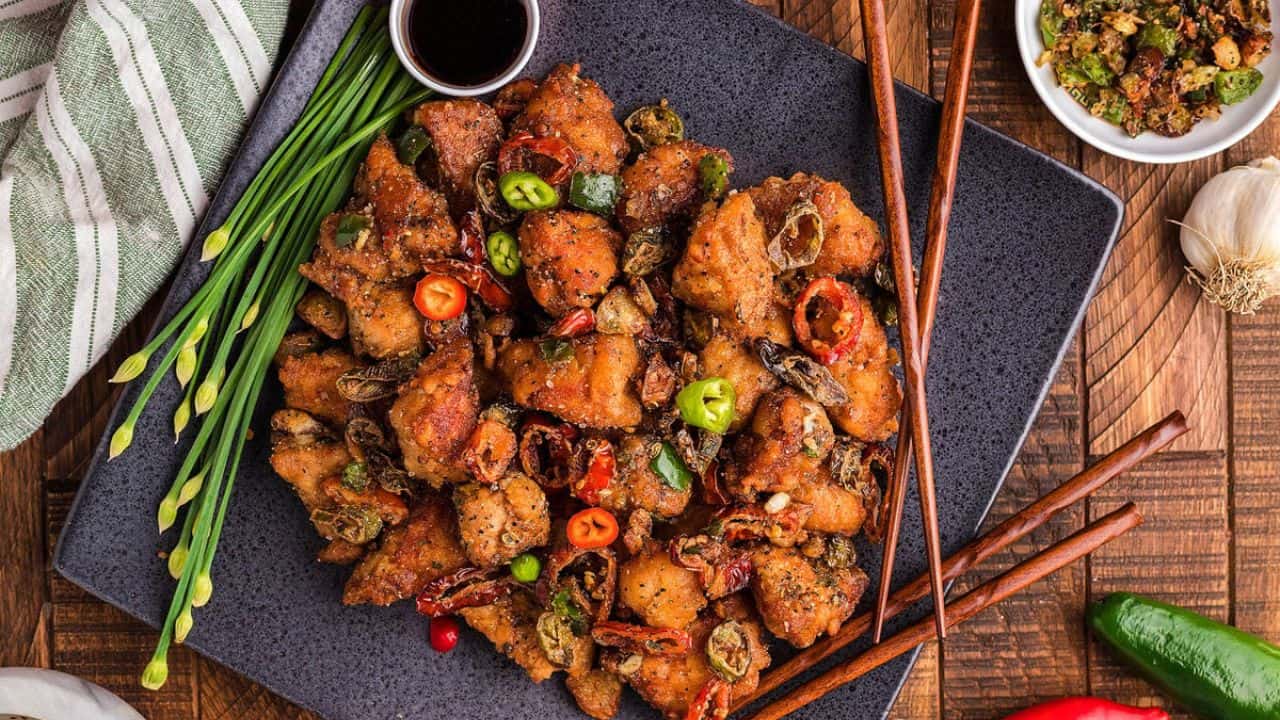 A plate of crispy fried chicken pieces garnished with sliced red and green chili peppers; chopsticks and a small bowl of dipping sauce are beside the plate.