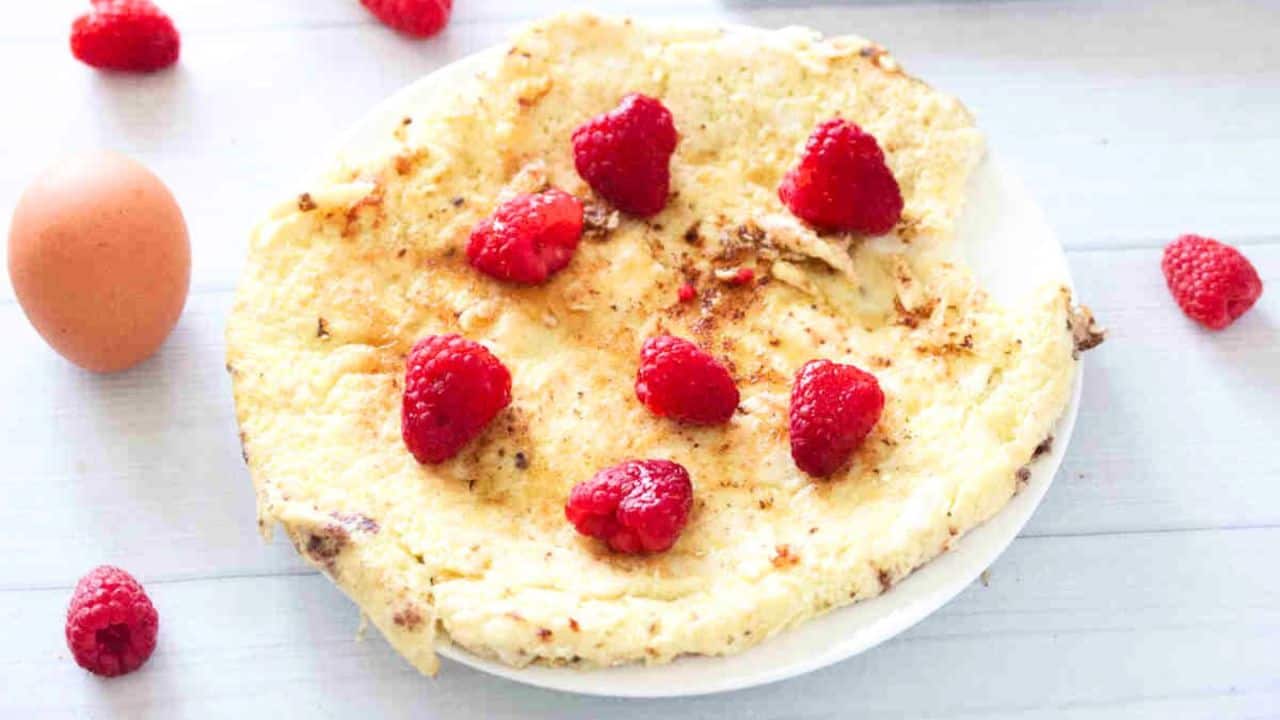 A plate with a cooked omelette topped with several fresh raspberries, placed on a light surface next to a whole egg and loose raspberries.
