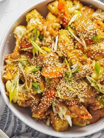 Bowl of stir-fried vegetables and tofu topped with sesame seeds and sliced green onions, served with a side of white rice.