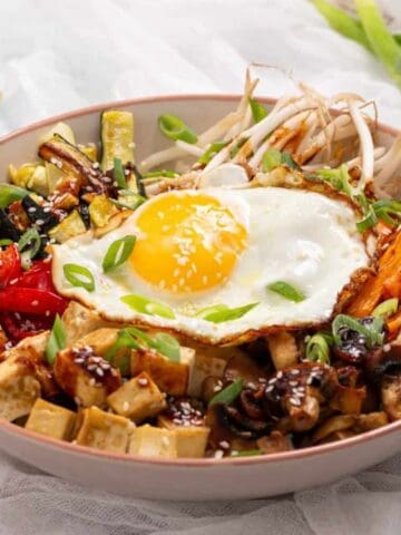 A bowl of rice topped with a sunny-side-up egg, tofu, sautéed vegetables, bean sprouts, and green onions, with a pair of chopsticks resting on the rim.