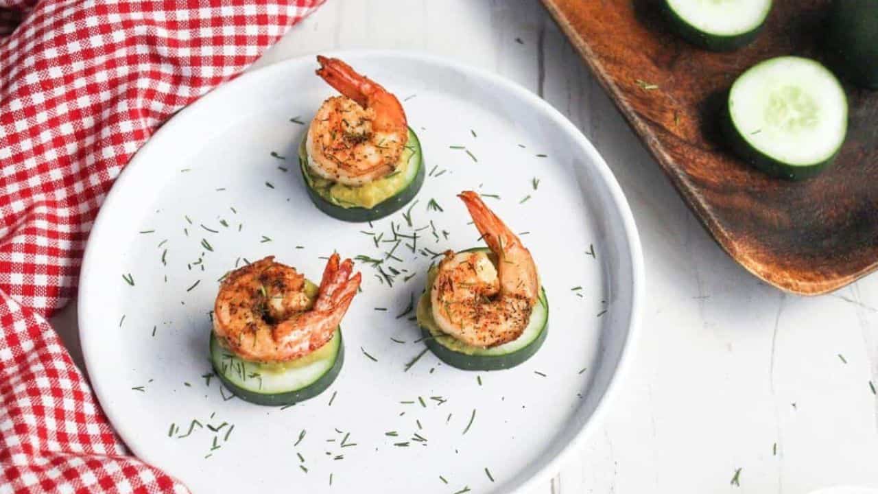 Three grilled shrimp atop cucumber slices garnished with herbs on a white plate with a red checkered napkin partially in view.