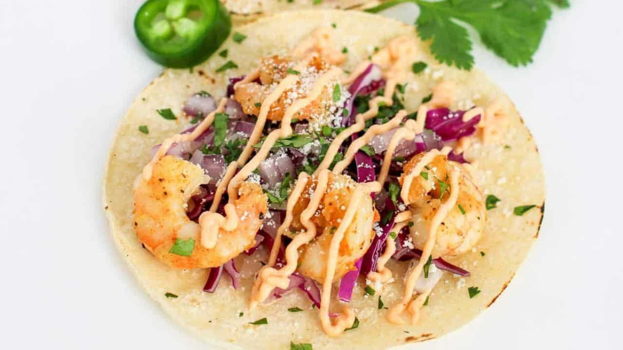 Shrimp taco with cabbage, onion, cilantro, sauce, cheese, and a lime wedge on a corn tortilla.