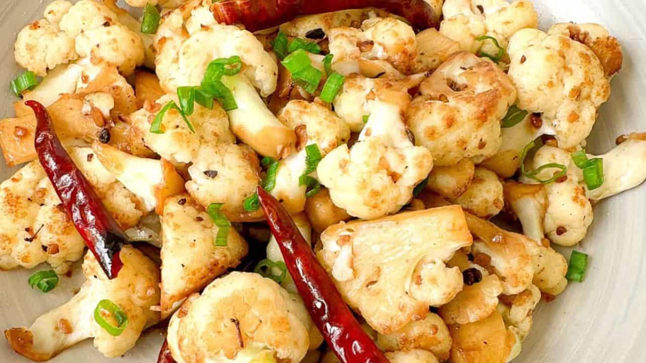 A bowl of stir fry Sichuan style cauliflower accompanied by red chili peppers and sliced green onions.