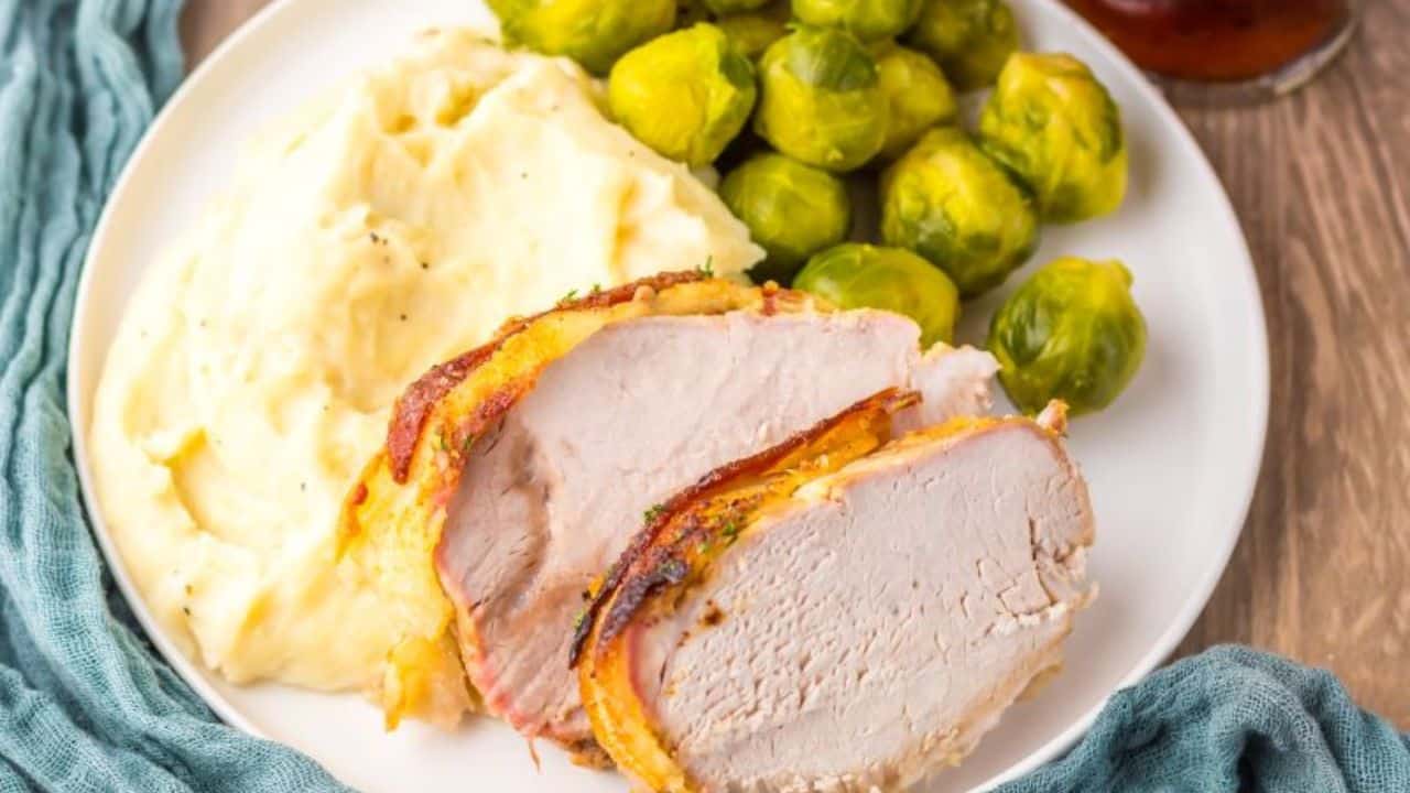 Plate with sliced roasted pork loin, mashed potatoes, and Brussels sprouts, set on a wooden table with a blue cloth and a glass of iced tea in the background.