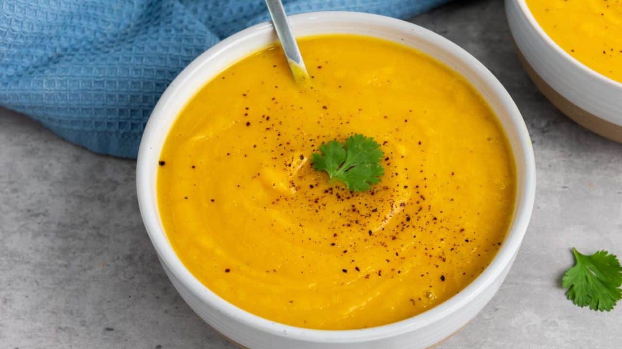 A bowl of creamy orange soup garnished with black pepper and a cilantro leaf, with a spoon resting inside the bowl. Another identical bowl is partially visible in the background.