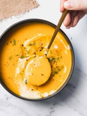 A hand holding a spoon over a bowl of creamy orange soup garnished with herbs, on a white marble surface.