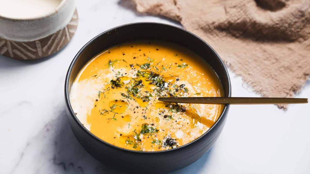 A black bowl filled with creamy orange soup, garnished with herbs, pepper, and a drizzle of oil. A gold spoon rests inside the bowl. A textured brown cloth and a cup are in the background.