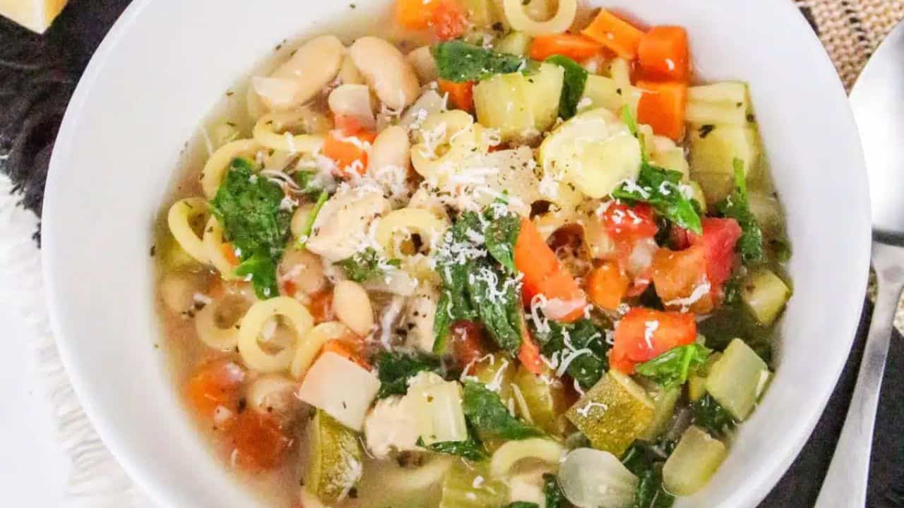 A white bowl of vegetable soup with pasta rings, carrots, zucchini, and greens, placed on a table with a spoon and carrot in the background.