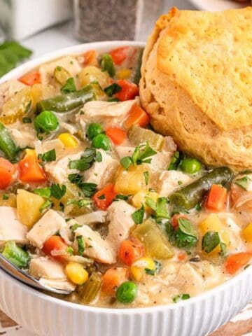 A bowl of creamy chicken and vegetable stew topped with a biscuit, with a spoon in the bowl and fresh parsley and a biscuit visible in the background.