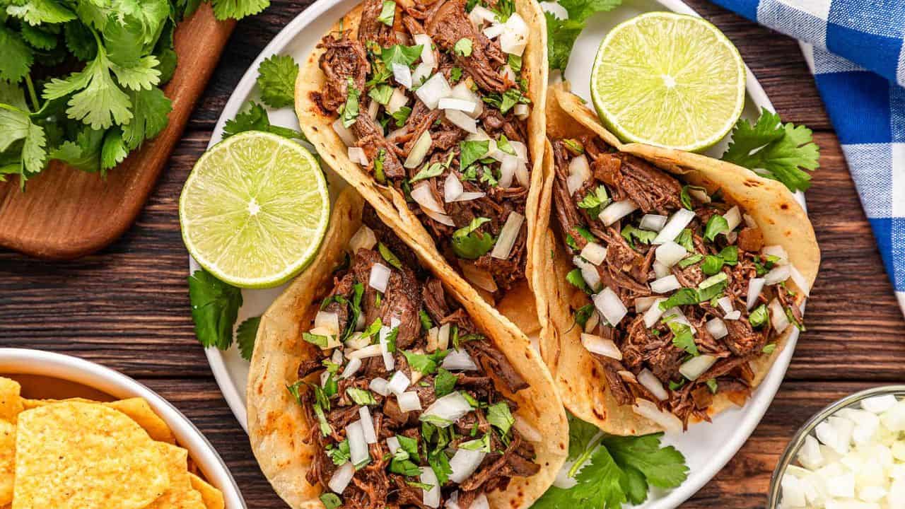 Three beef tacos topped with chopped onions and cilantro are served on a white plate with lime halves, surrounded by fresh cilantro and tortilla chips.