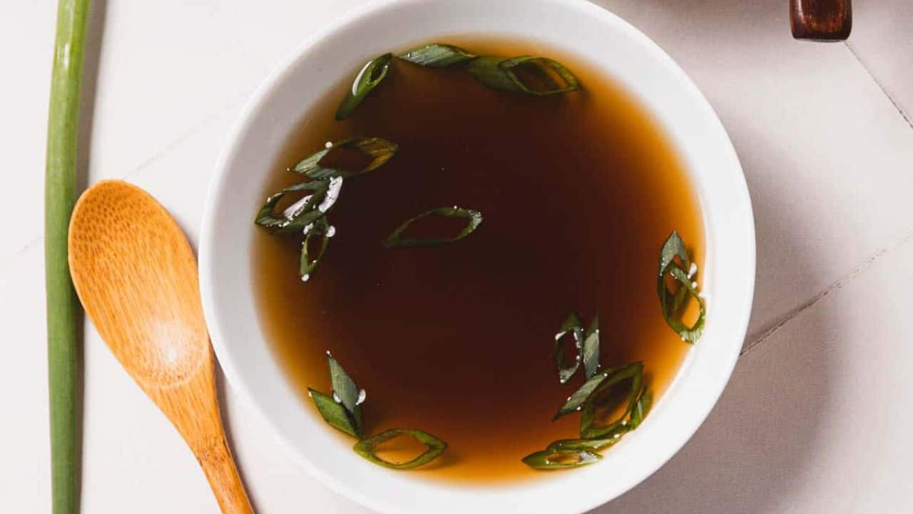 Two white bowls filled with a dark broth garnished with sliced green onions. Wooden spoons are placed beside each bowl. Green onion stalks are scattered on a light tiled surface.