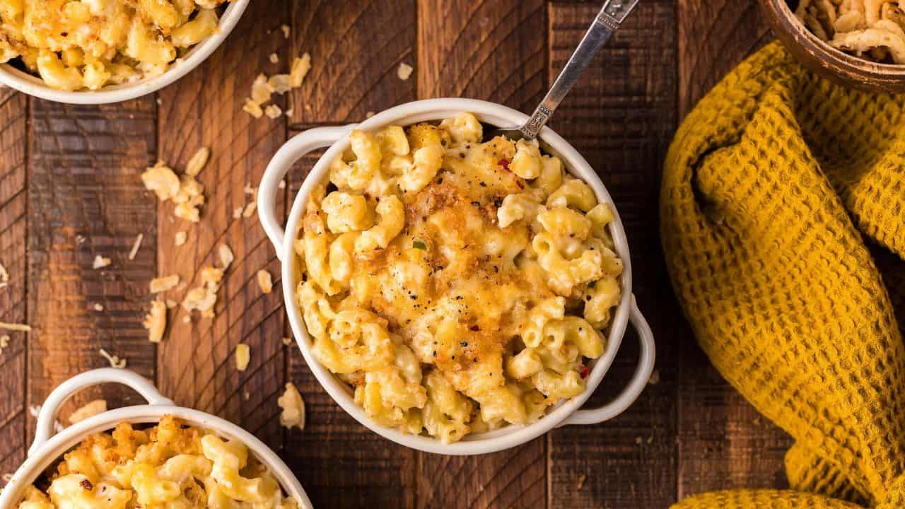 Baked macaroni and cheese in white ceramic bowls on a wooden surface, topped with a golden crust and served with a spoon; a yellow cloth napkin is beside the bowls.