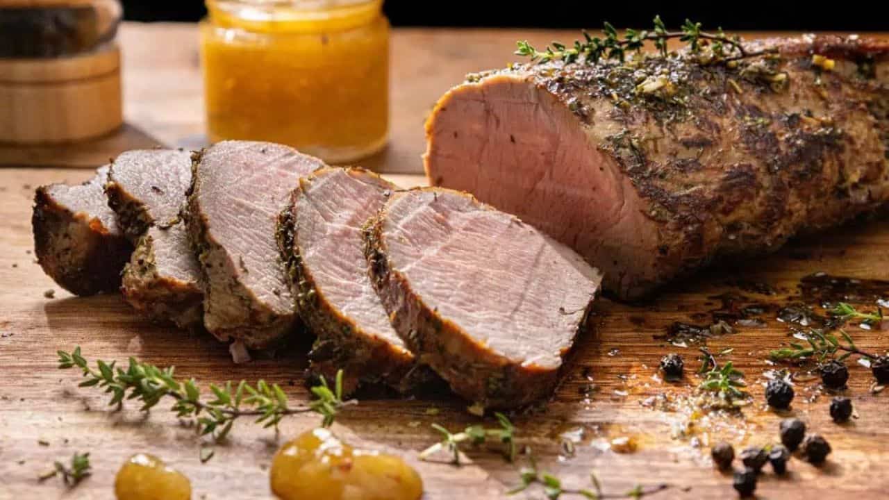 Slices of roasted pork tenderloin on a wooden board, garnished with fresh herbs. A jar of mustard is in the background, along with peppercorns scattered around the meat. The scene is warmly lit and appetizing.