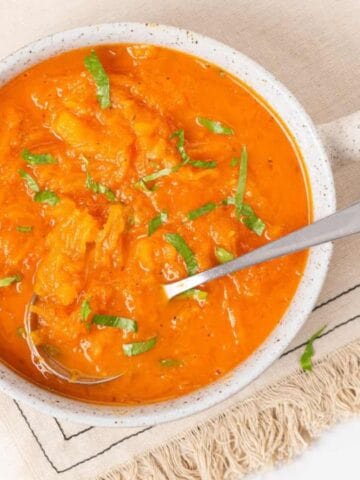 A bowl of orange soup garnished with chopped herbs sits on a beige cloth napkin, with a spoon resting inside the bowl.