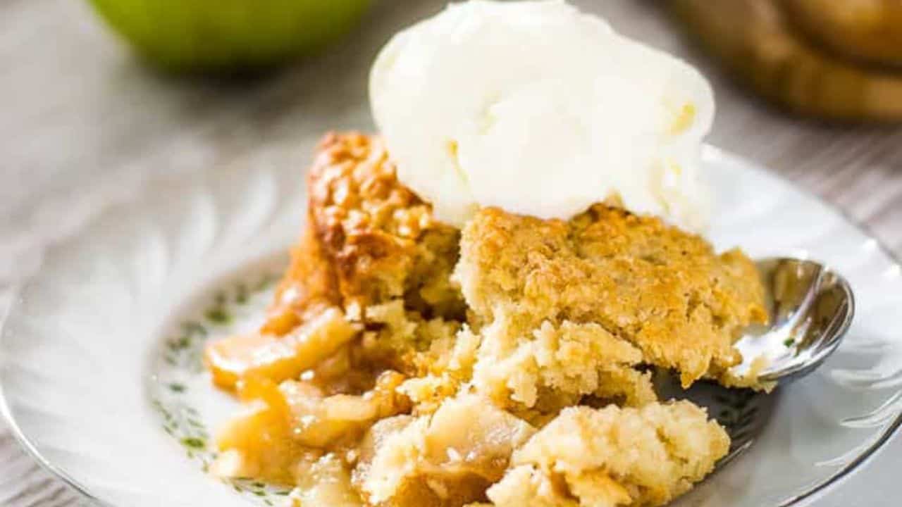 A spoonful of oatmeal with diced apples hovers above a white bowl filled with creamy oatmeal. A green pear and a cinnamon stick tempt in the background, hinting at cheat day desserts that delight without breaking rules.