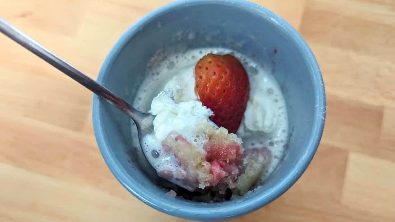 A blue cup with whipped cream, a whole strawberry, and a metal spoon inside. The cup is on a wooden surface.