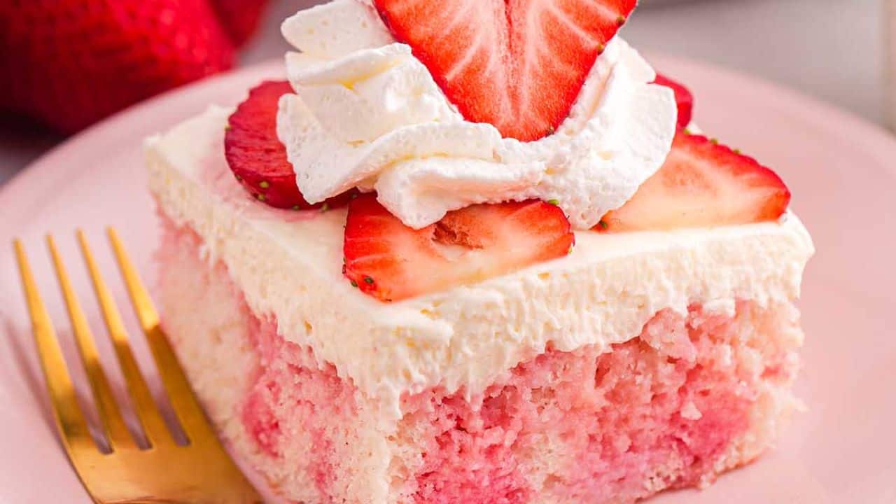 A slice of strawberry poke cake topped with whipped cream and fresh strawberry slices on a pink plate with a gold fork.