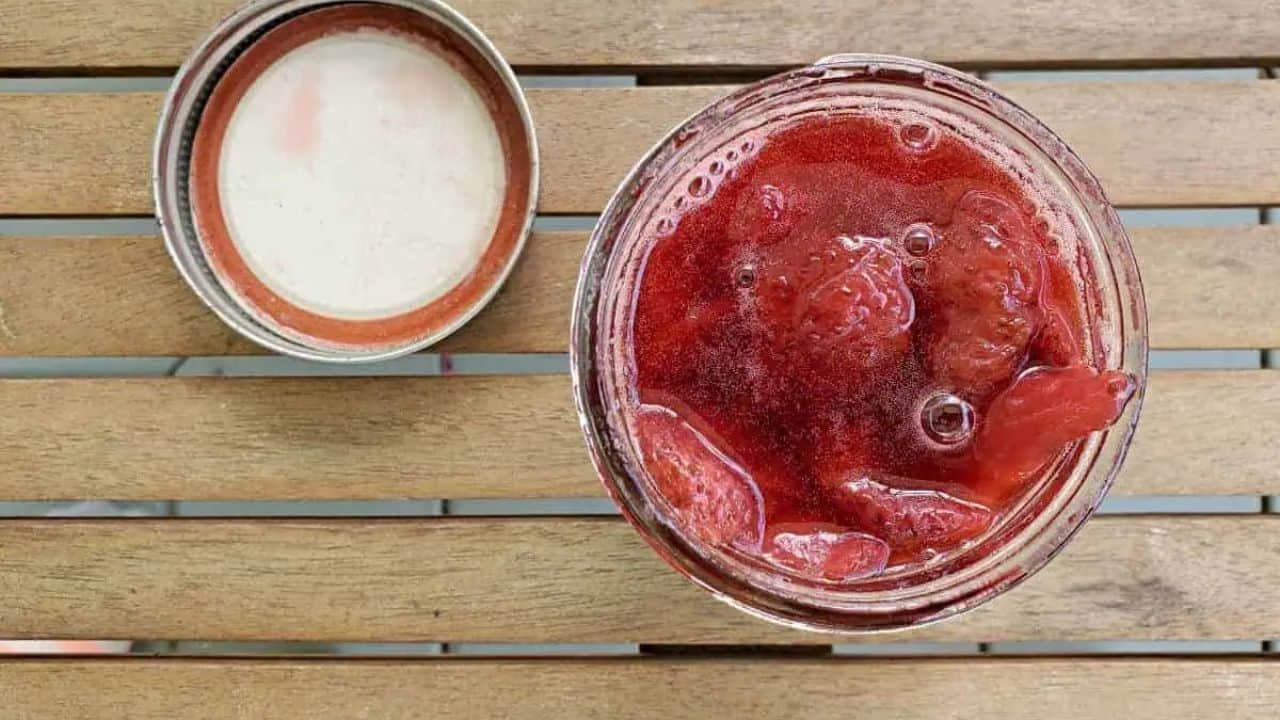 Spoon adding strawberry syrup to a glass of ice cream with a jar of strawberry syrup behind it.