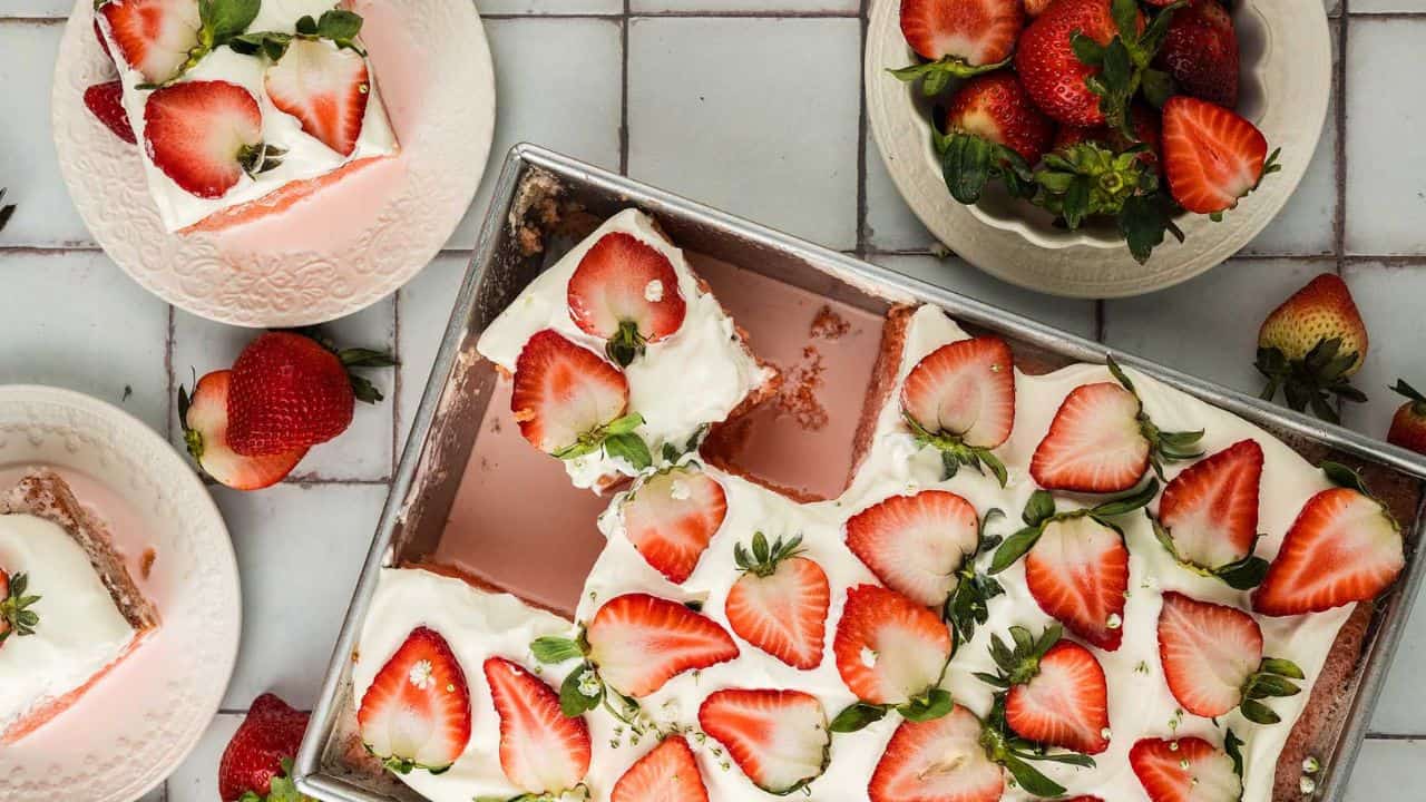 A frosted sheet cake topped with sliced strawberries is cut into squares, with whole strawberries and cake pieces on plates nearby.
