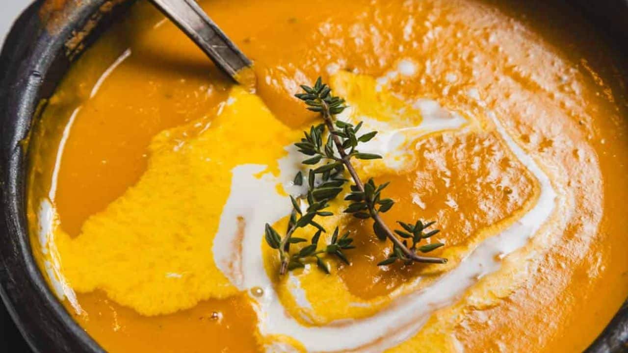 A bowl of creamy orange soup is placed on a black plate, garnished with a swirl of cream and a sprig of herb. A spoon rests inside the bowl. Small sprigs of thyme are scattered on the marble surface nearby.