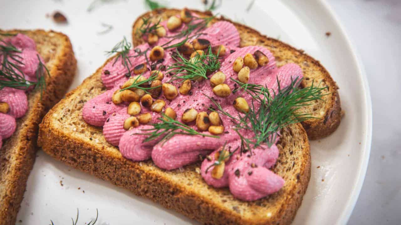 Two slices of whole grain toast topped with piped pink spread, toasted pine nuts, and fresh dill, served on a white plate.