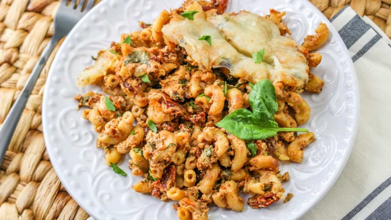 A plate of macaroni with sun-dried tomatoes and a melted cheese topping, garnished with basil, on a woven placemat.