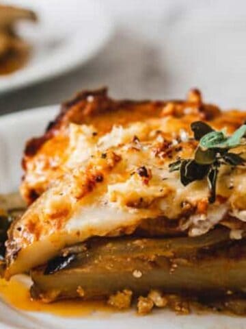 A serving of baked moussaka with layers of eggplant, meat sauce, and béchamel, garnished with fresh herbs, on a white plate.