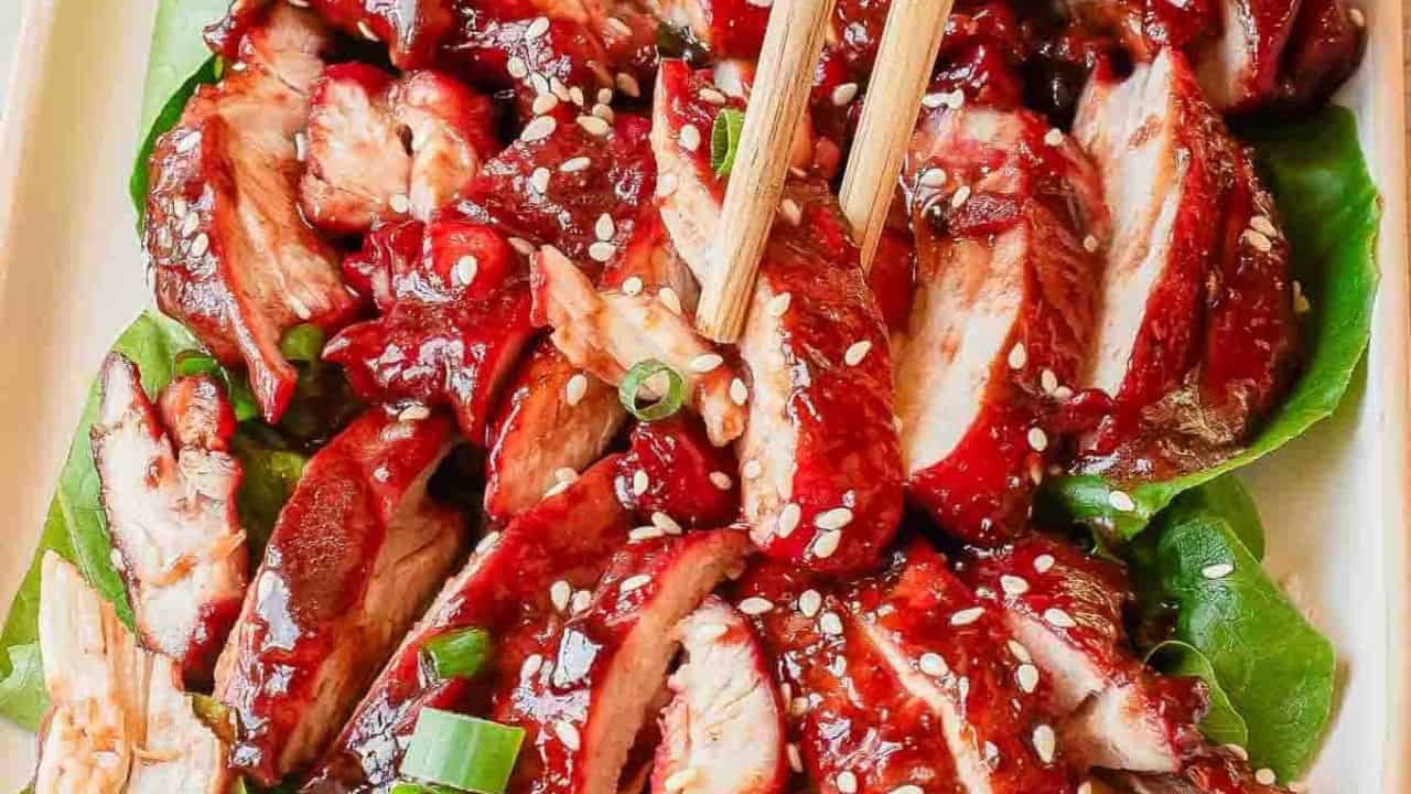A bowl of white rice topped with sliced red-glazed meat, chopped green onions, and white sesame seeds, with chopsticks picking up a piece.