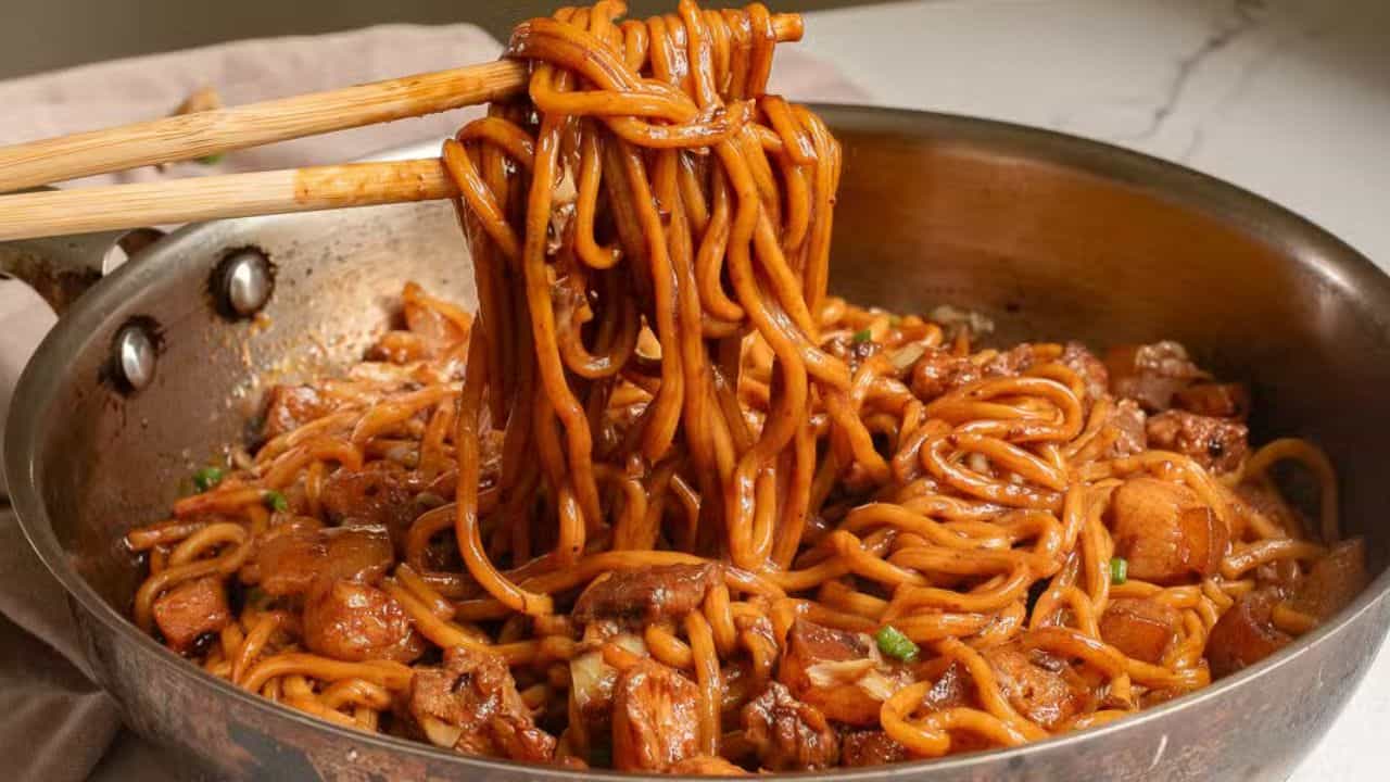 A close-up of a pan filled with savory noodles and chunks of meat being lifted by chopsticks. The dish appears well-seasoned with sauce, and green garnishes are sprinkled on top. A beige napkin and a piece of dumpling are in the background.