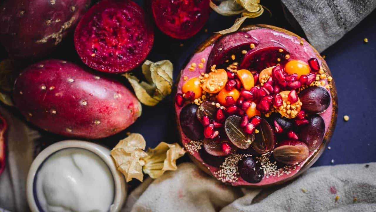 A smoothie bowl topped with halved grapes, sliced plums, pomegranate seeds, golden berries, chia seeds, and bee pollen, placed next to a vintage spoon on a beige cloth.