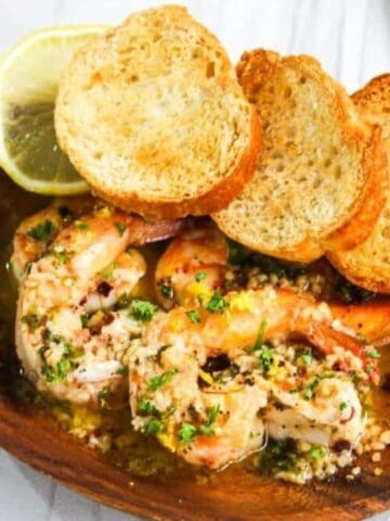 Wooden plate with cooked shrimp in garlic sauce, topped with herbs and crumbs, served with lemon slice and three pieces of toasted bread.