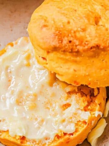 A split biscuit on a plate, with one half spread with butter and a knife resting beside it.