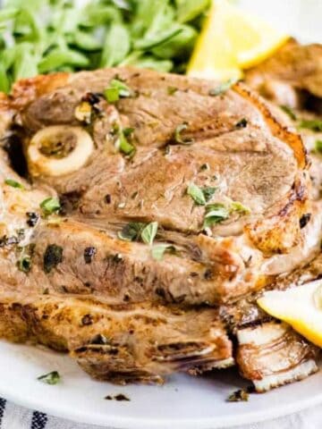 Grilled lamb chop garnished with herbs, served with arugula and lemon wedges on a white plate.