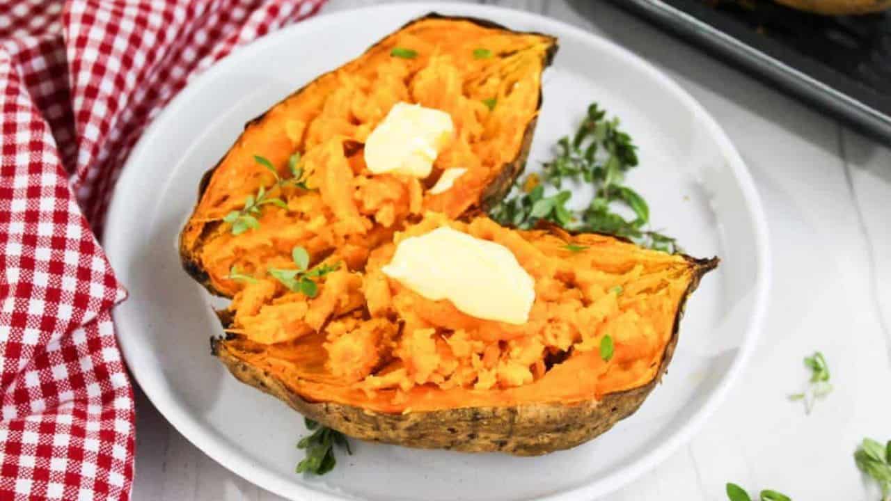 Two halves of baked sweet potato topped with butter and garnished with herbs on a white plate, with a red checkered napkin on the side.
