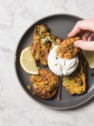 A hand dips a vegetable fritter into a dollop of yogurt on a gray plate, garnished with lemon slices, atop a marble countertop.