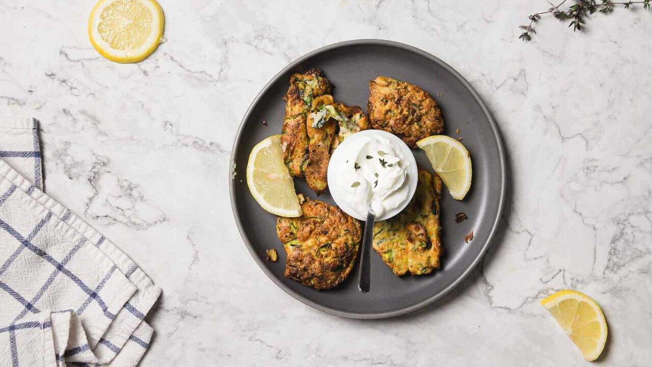 A hand dips a fried zucchini fritter into a small bowl of white cream sauce on a gray plate. Other fritters and lemon wedges are arranged around the bowl on a marble surface.