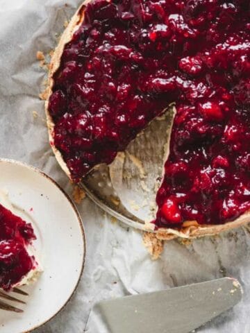 A cherry pie with a slice removed sits on parchment paper; the slice is served on a small plate with a fork beside it.