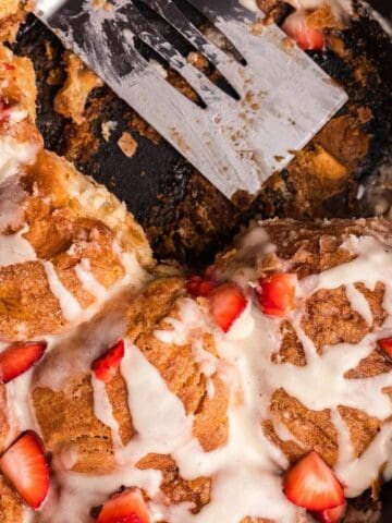 A skillet of cinnamon rolls topped with icing and fresh strawberry pieces, with a metal spatula resting inside and a red and white towel nearby.