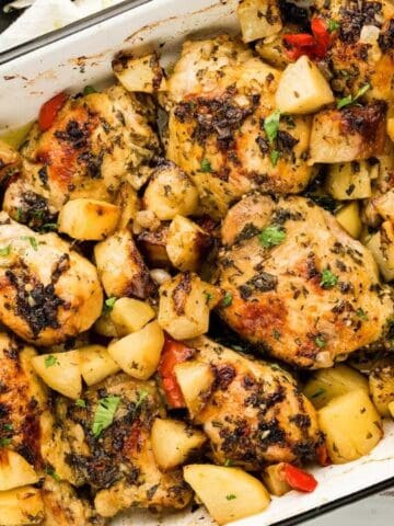 Baked chicken thighs with roasted potatoes and red peppers in a white rectangular baking dish, garnished with herbs.