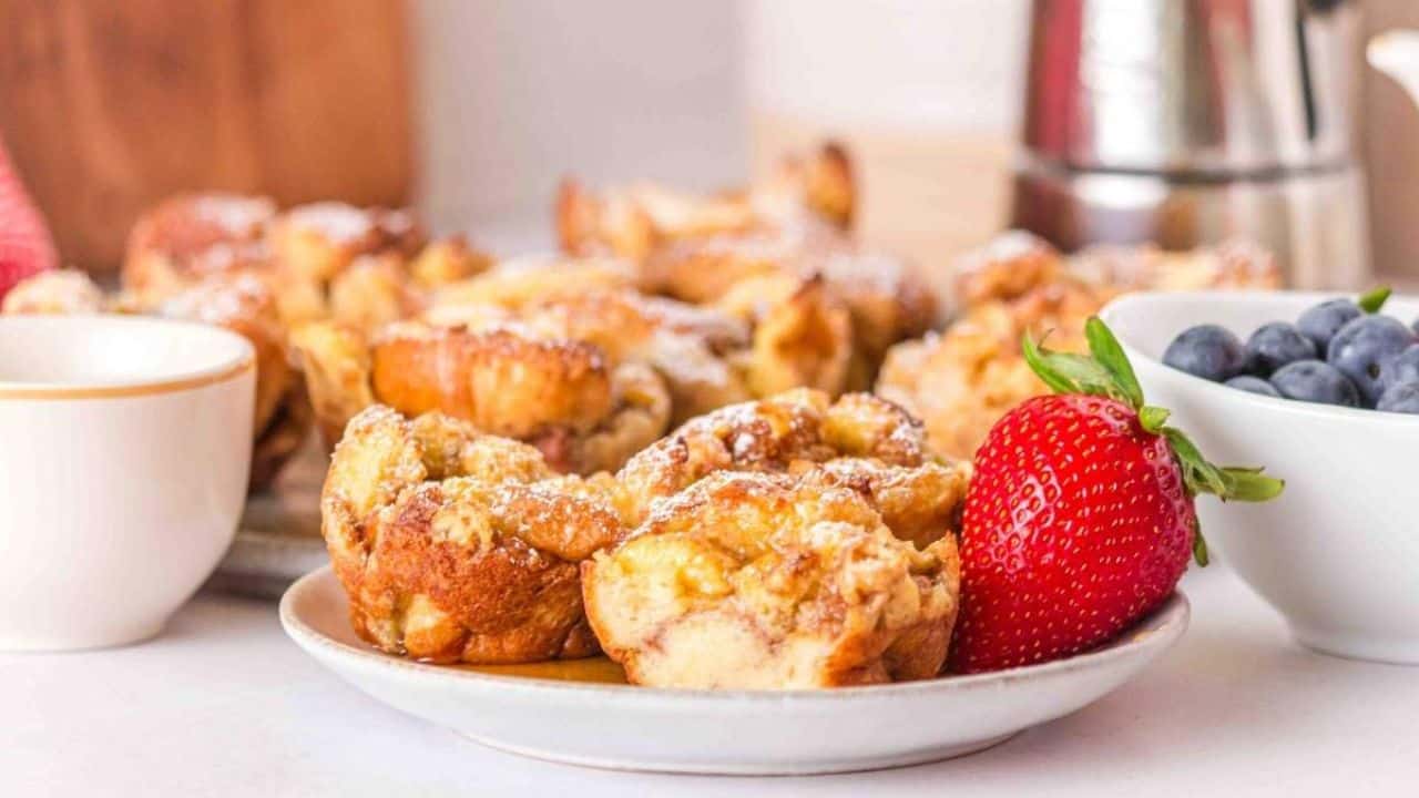 A plate of cinnamon roll muffins dusted with powdered sugar, served with a fresh strawberry and a bowl of blueberries on a white table.