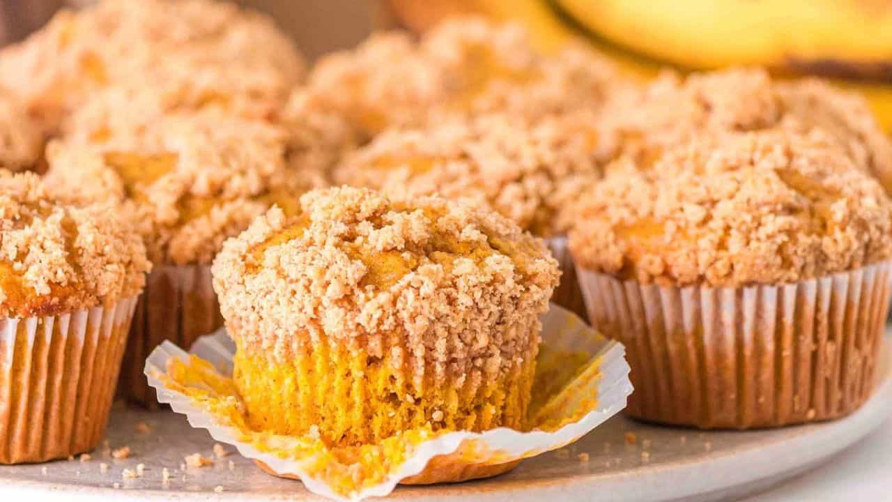 Several pumpkin muffins with crumb topping are displayed on a light-colored surface, with one muffin partially unwrapped in the foreground.