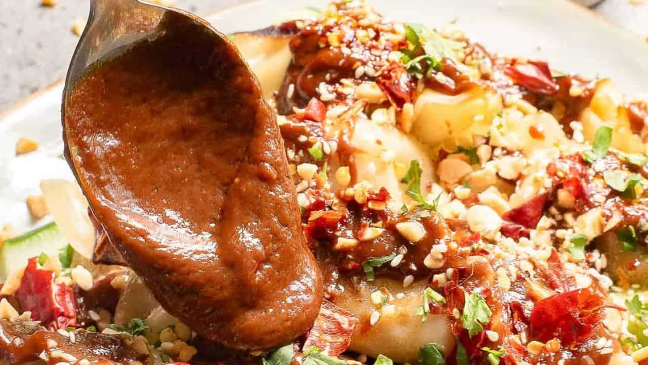 A spoon drizzles rich peanut sauce over dumplings topped with chopped peanuts, red chili pieces, and fresh herbs on a plate. The dish appears appetizing and flavorful, highlighting the combination of textures and colors.