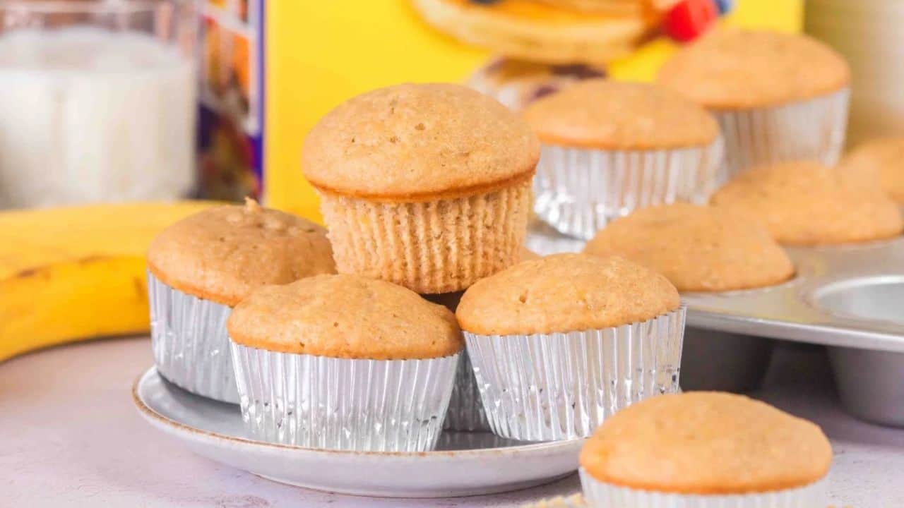 Banana muffins in silver foil cups are stacked on a plate, with a banana and a glass of milk in the background.