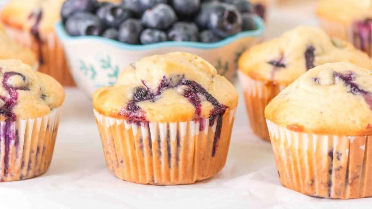 Several blueberry muffins are arranged on a surface with a bowl of fresh blueberries in the background.