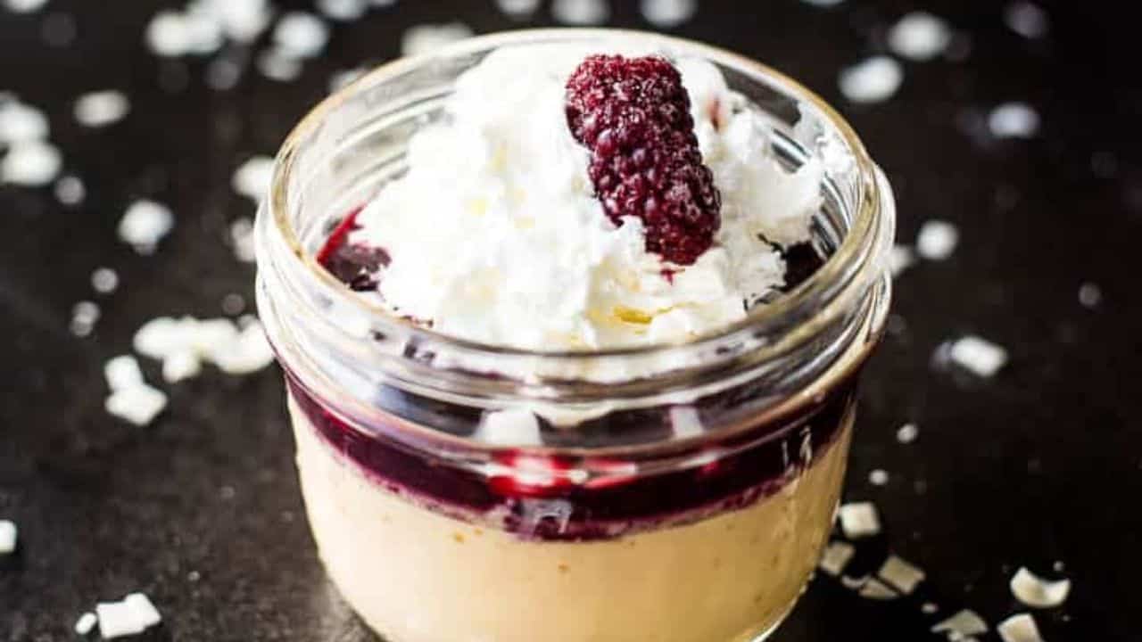 Three blackberry pots de creme lined up on a wooden board next to silver spoons.
