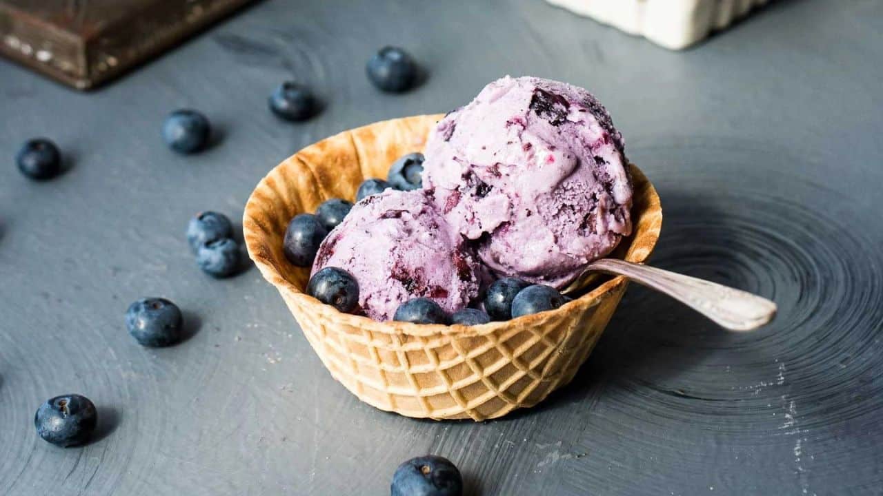 An overhead shot of homemade blueberry ice cream in a metal loaf pan next to waffle bowls, spoons, and a cream colored napkin.