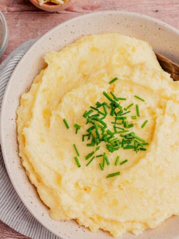 A bowl of mashed potatoes topped with chopped chives, with a wooden spoon resting inside, placed on a cloth napkin on a wooden table.