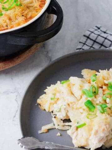 A serving of cheesy hashbrown casserole garnished with chopped green onions on a gray plate, with the remaining casserole in a black dish nearby.