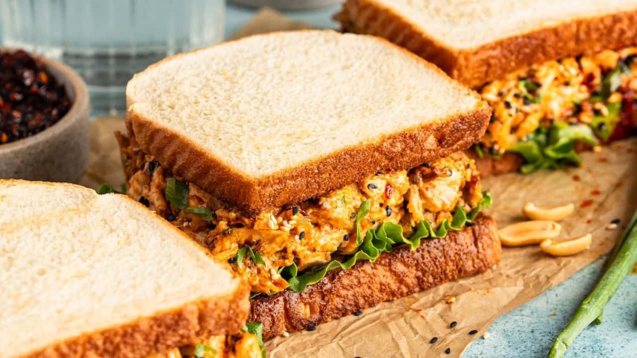 Three sandwiches filled with a creamy, mixed chicken salad and leafy lettuce are arranged on parchment paper, with a bowl of dark spices and peanuts nearby.
