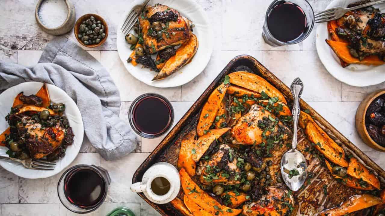 Baked chicken with sweet potatoes, olives, and herbs served on plates and a tray, accompanied by glasses of red wine and small bowls of condiments on a light table.